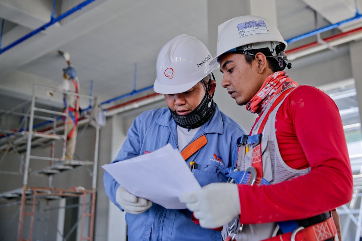 Two construction workers in safety gear reviewing blueprints at a work site.