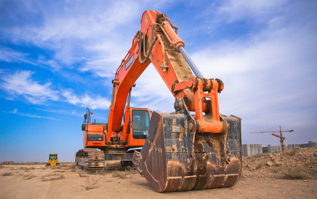 Orange excavator on a construction site under a blue sky, with cranes in the background.