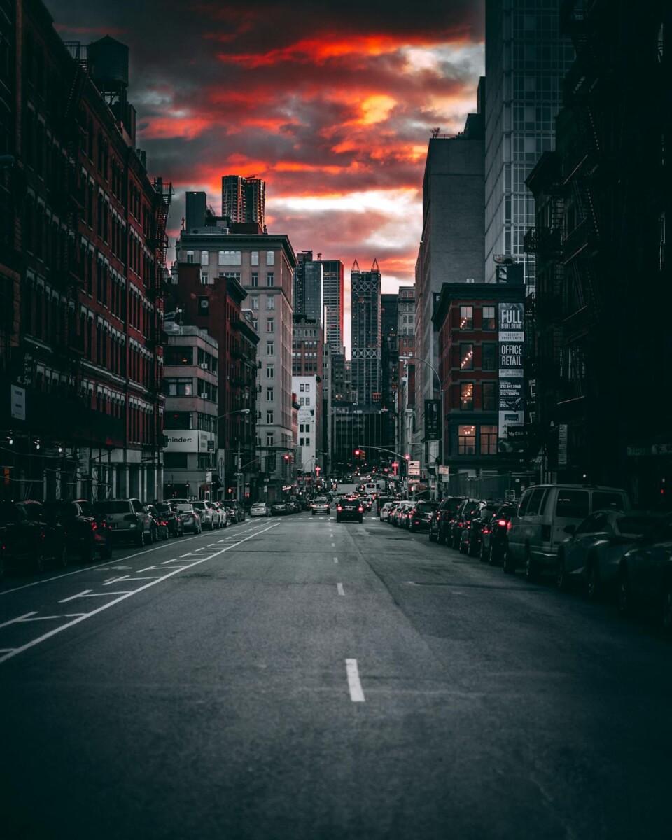 City street at dusk, lined with buildings, beneath a dramatic red and gray clouded sky.