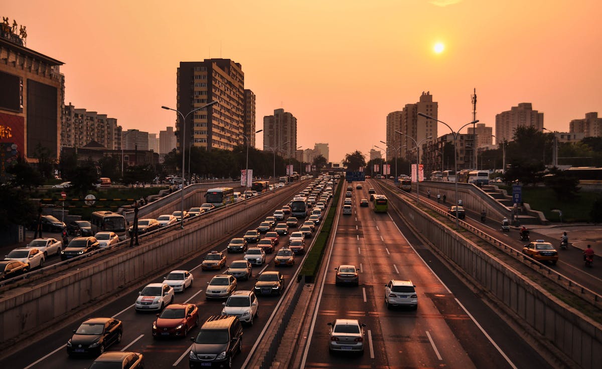 Traffic on a multi-lane highway at sunset, surrounded by city buildings under an orange sky.