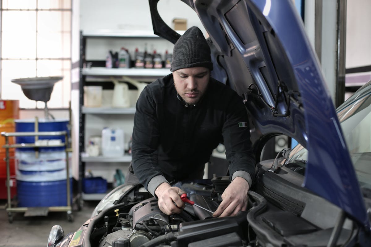 Mechanic wearing beanie repairs engine under open hood in a garage.