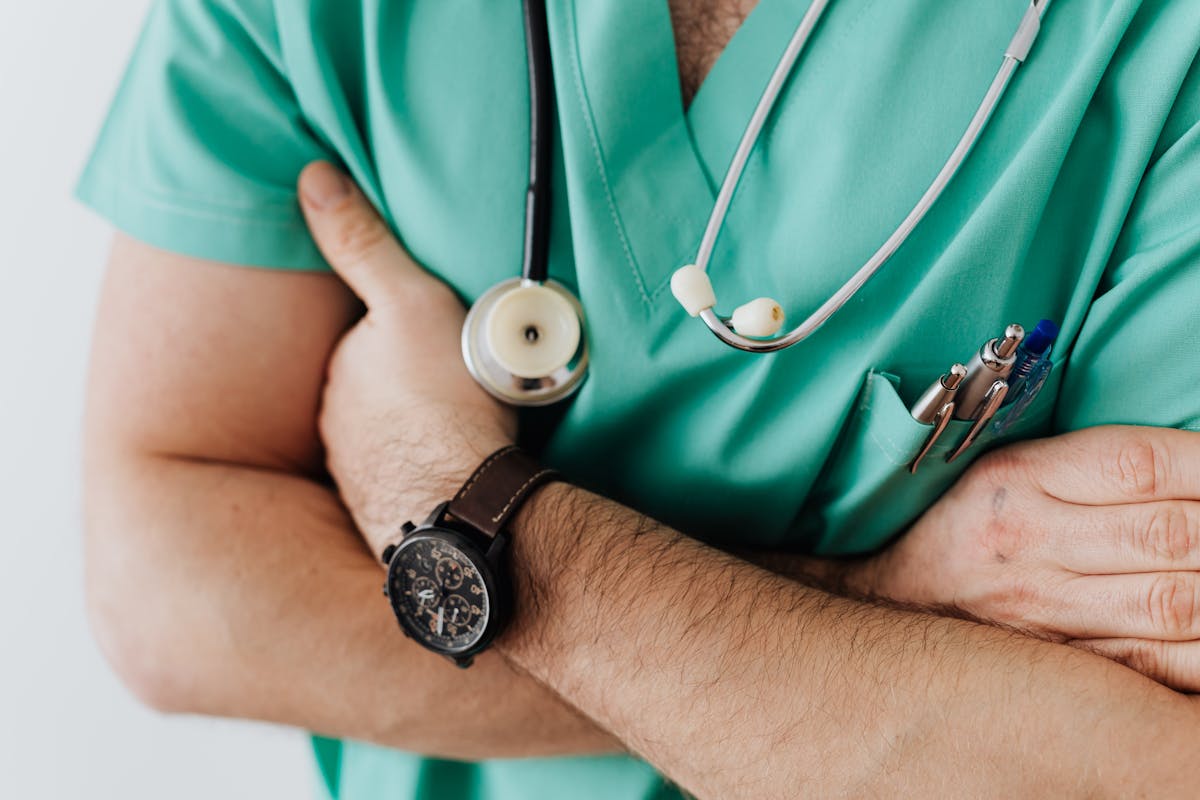 Doctor in green scrubs with stethoscope and arms crossed, wearing a watch and pens in the pocket.