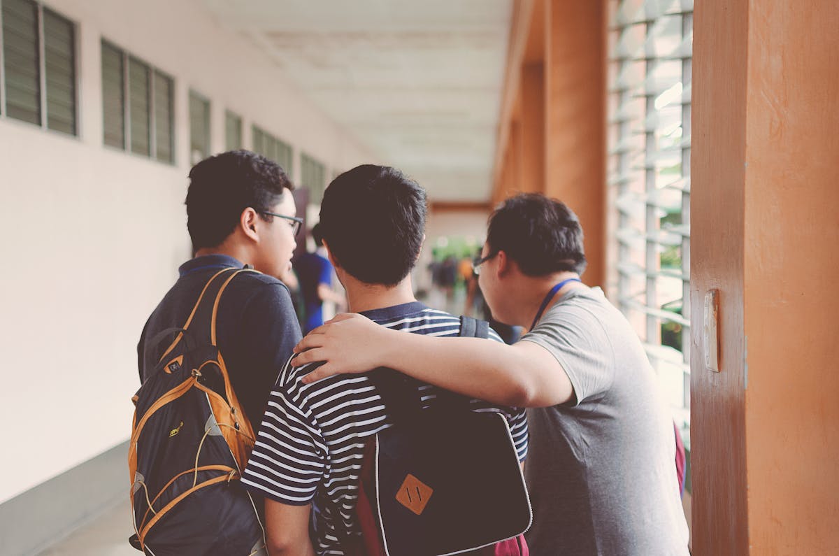Three friends walking down a hallway, one with an arm around another, wearing backpacks.