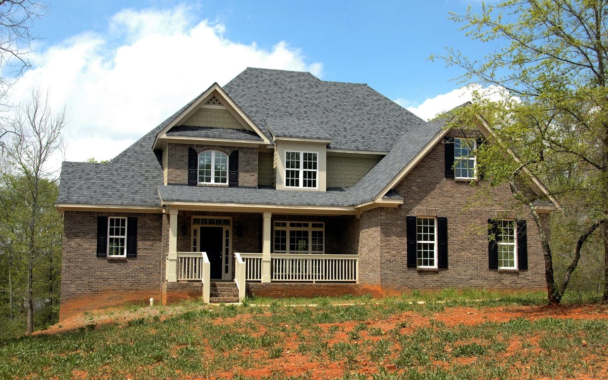 Two-story brick house with gray gabled roof, white trim, and a front porch, set on a grassy area with trees around.