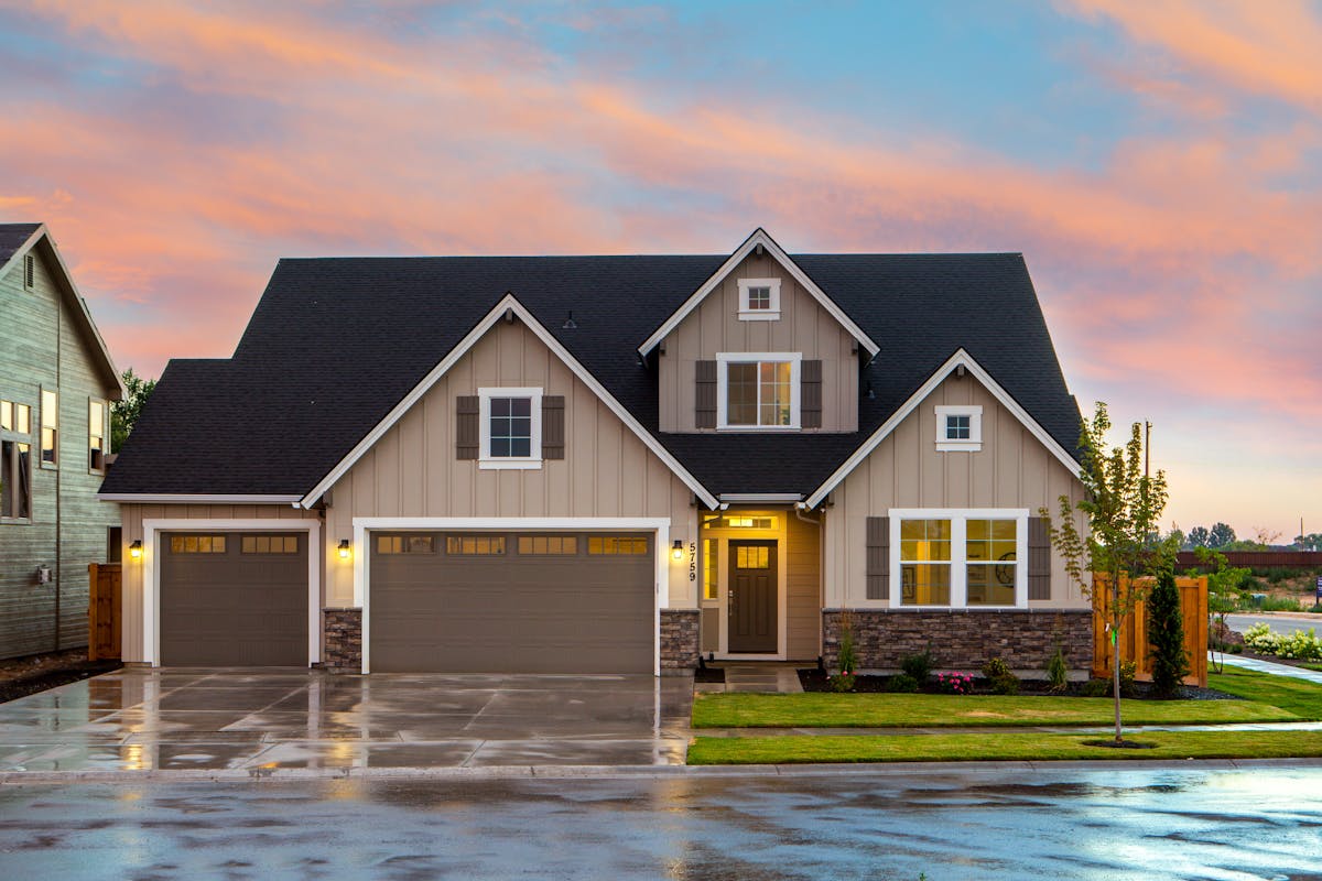 Modern suburban house with a double garage, lit windows, and a wet driveway under a colorful sunset sky.
