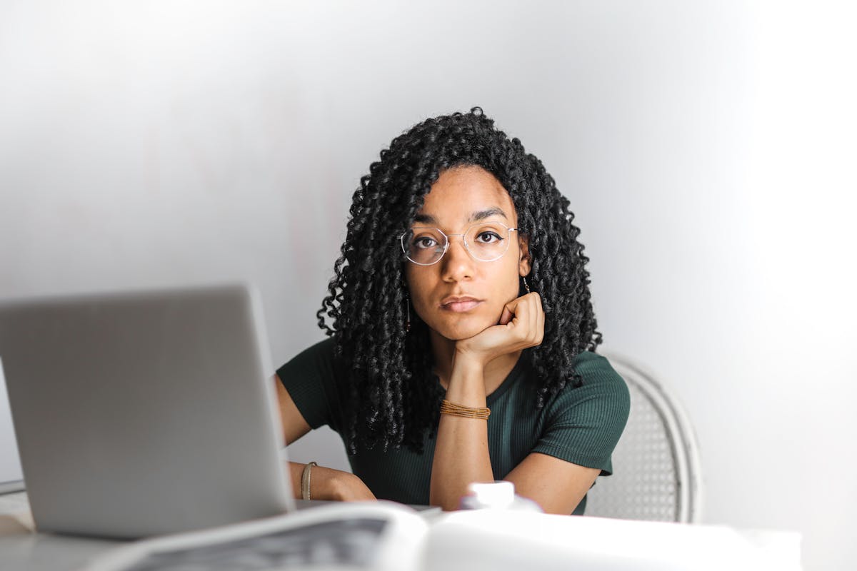 Person with glasses sitting at a desk with a laptop, resting chin on hand, looking thoughtful.